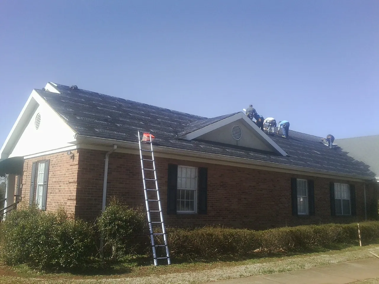 Crew tearing shingles off a brick ranch roof with a ladder set — Emergency Roof Repair work by our crew in Louvale
