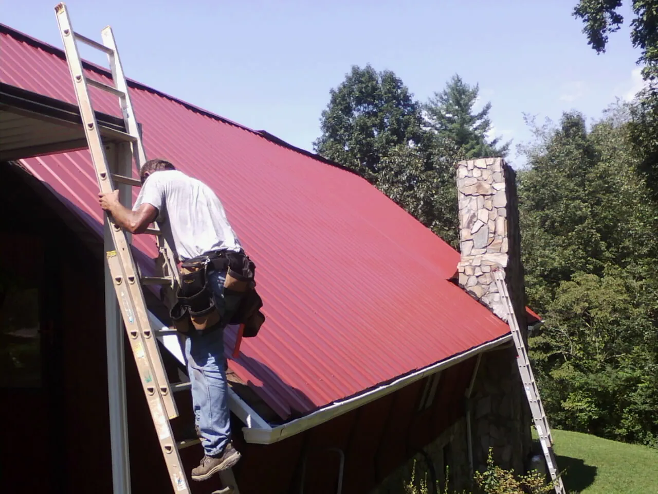 Roofer on a ladder beside a red metal roof and stone chimney — Soffit & Fascia Repair by Roofing Brothers in Louvale